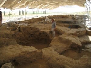 A white man stands in an archaeological excavation with a canvas shelter overhead
